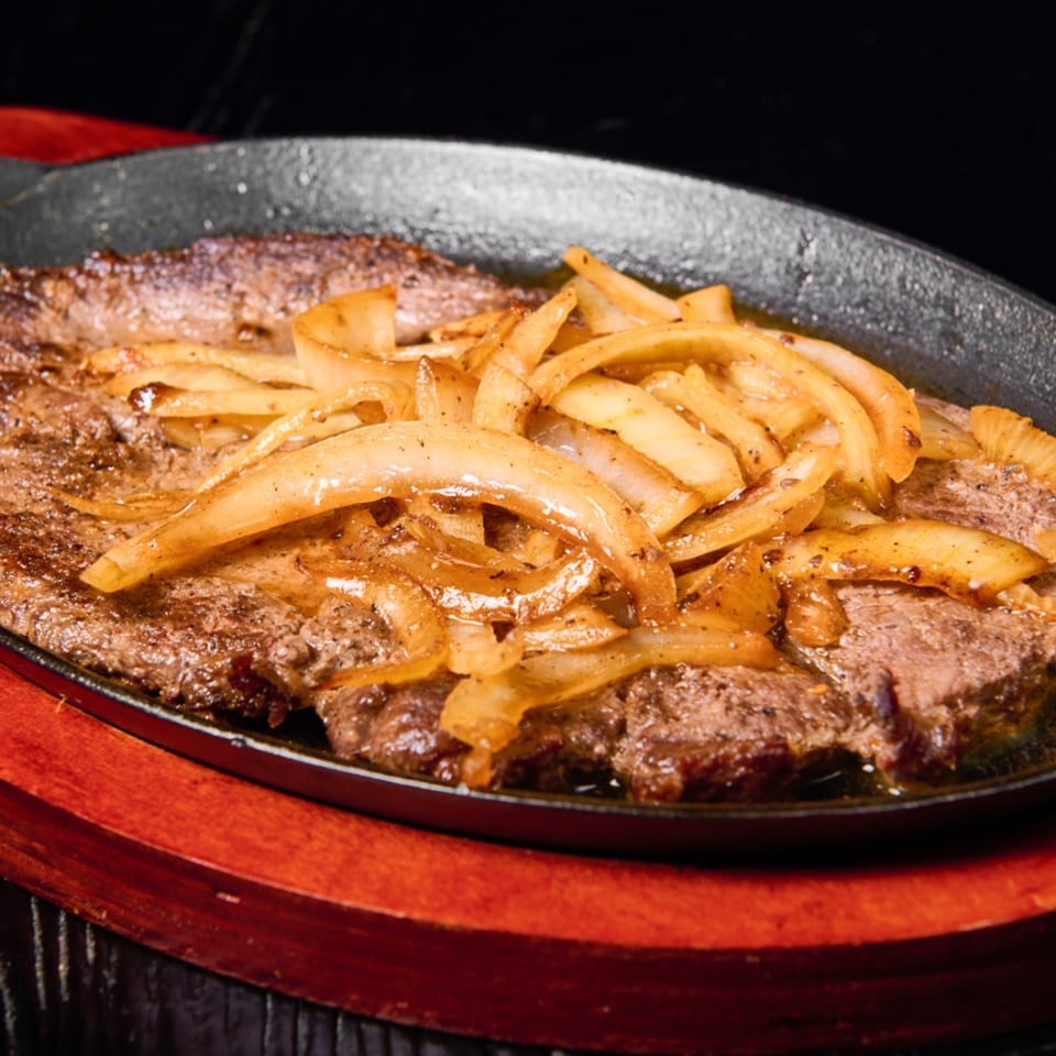Sizzling steak topped with caramelized onions in a black skillet, served on a wooden base.