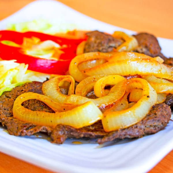 Dominican beef steak with sautéed onions, red peppers, and shredded lettuce on a white plate.