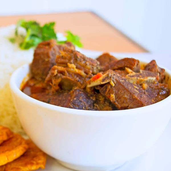 A white bowl filled with a rich, dark stew, accompanied by white rice and plantains.