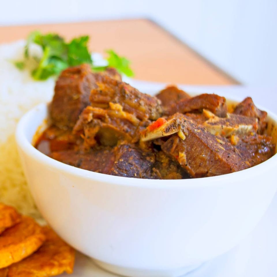A white bowl filled with a rich, dark stew, accompanied by white rice and plantains.